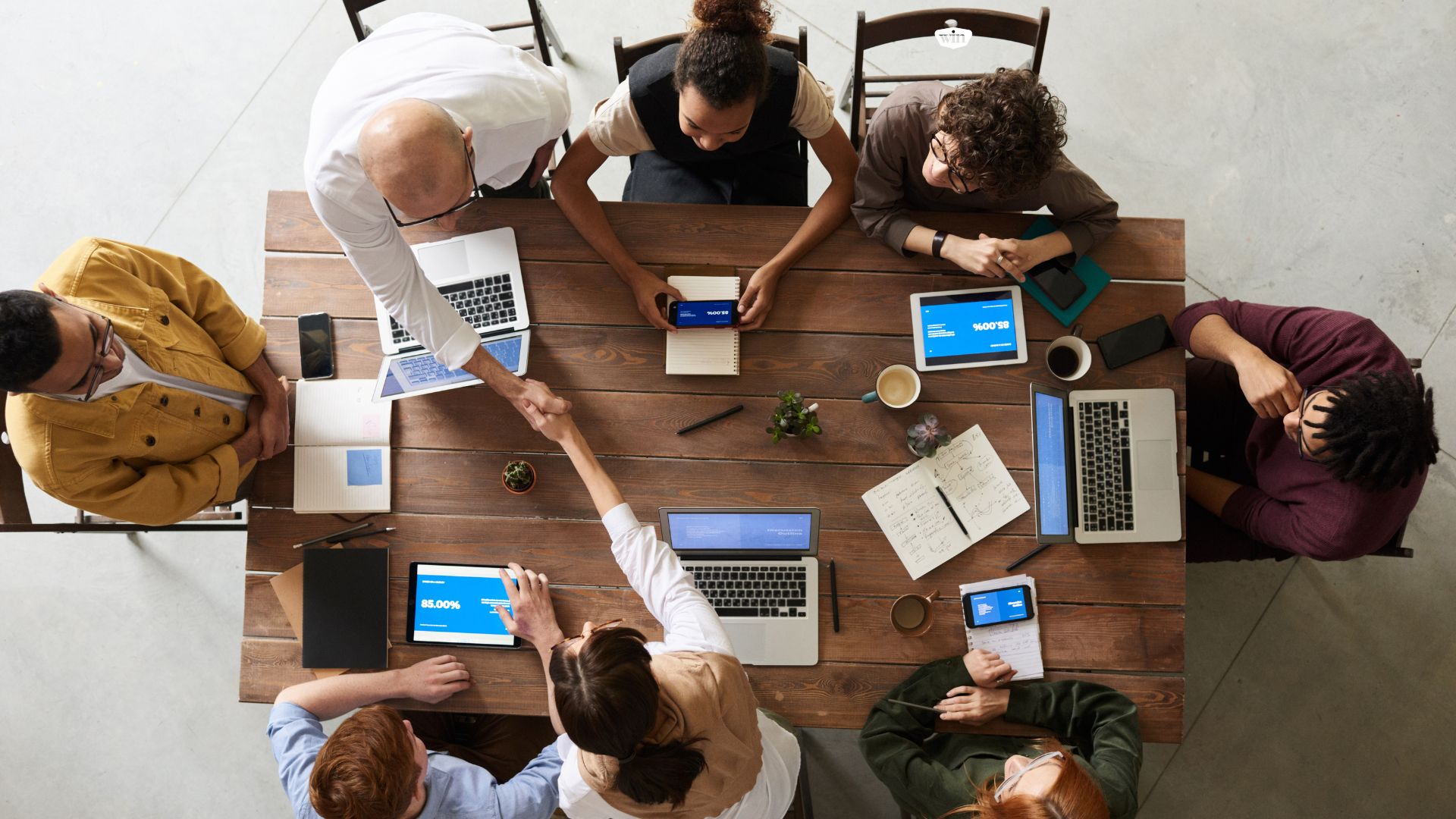 Team meeting around a table working on their endpoint devices. Two professionals are shaking hands across the table.